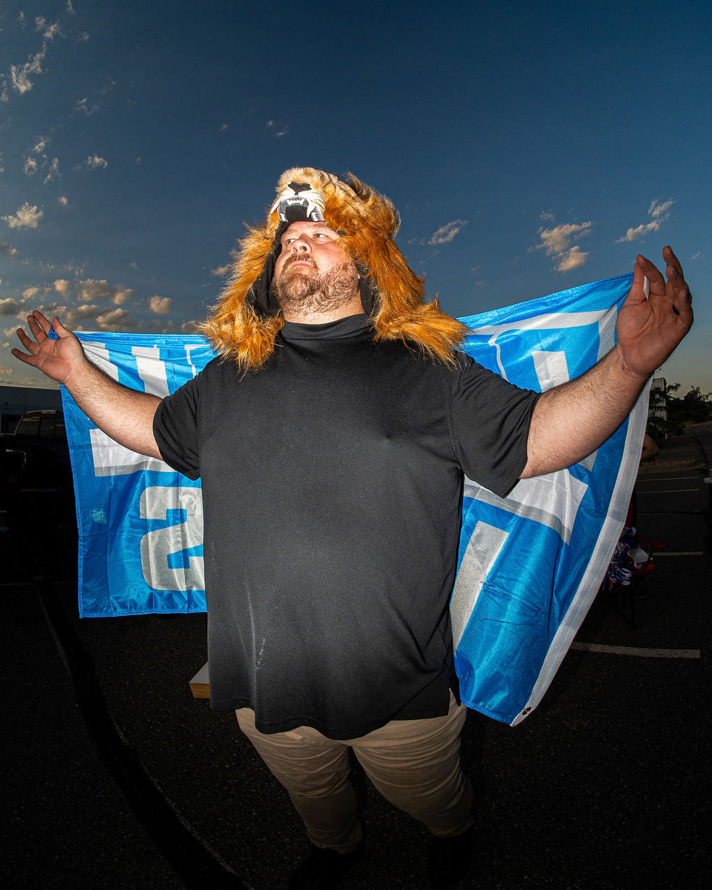 A man wearing a lion costume headpiece and a black t-shirt stands outdoors with his arms outstretched, holding a blue and white flag against a dark sky background.