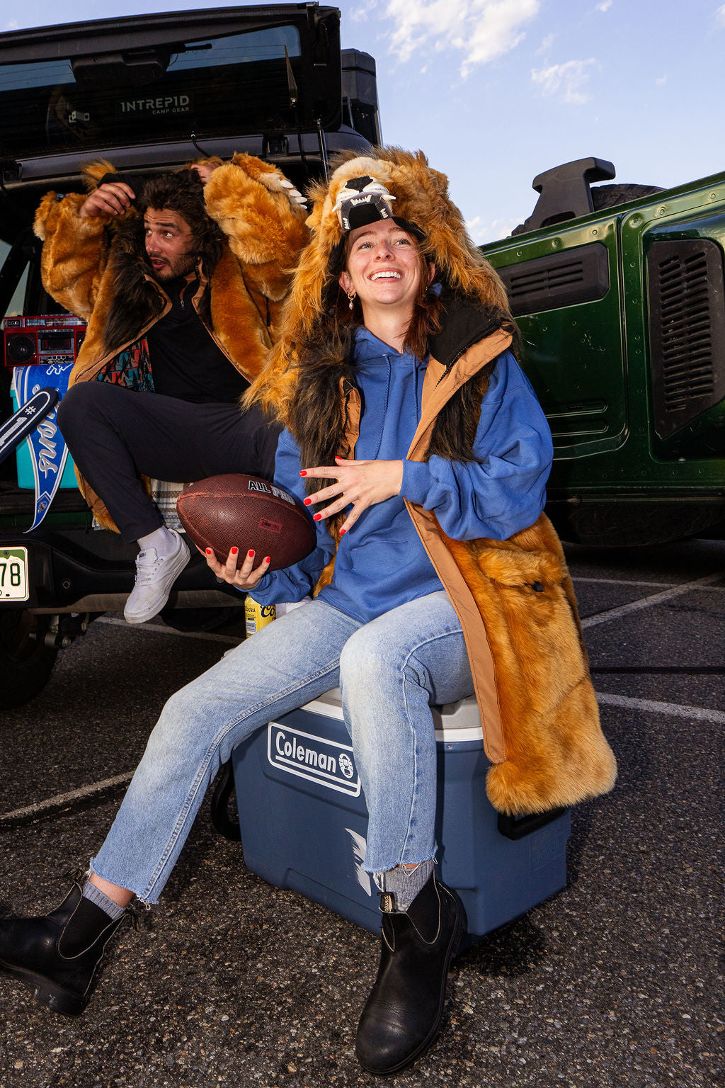 Two people outdoors in front of vehicles, wearing wildcoat lion parkas. The person in the foreground sits on a blue Coleman cooler holding a football, while another person is seated behind on the back of a black vehicle.