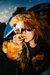 A person wearing a large lion costume headpiece and sunglasses, eating a hot dog outdoors in front of a vehicle under a blue sky.
