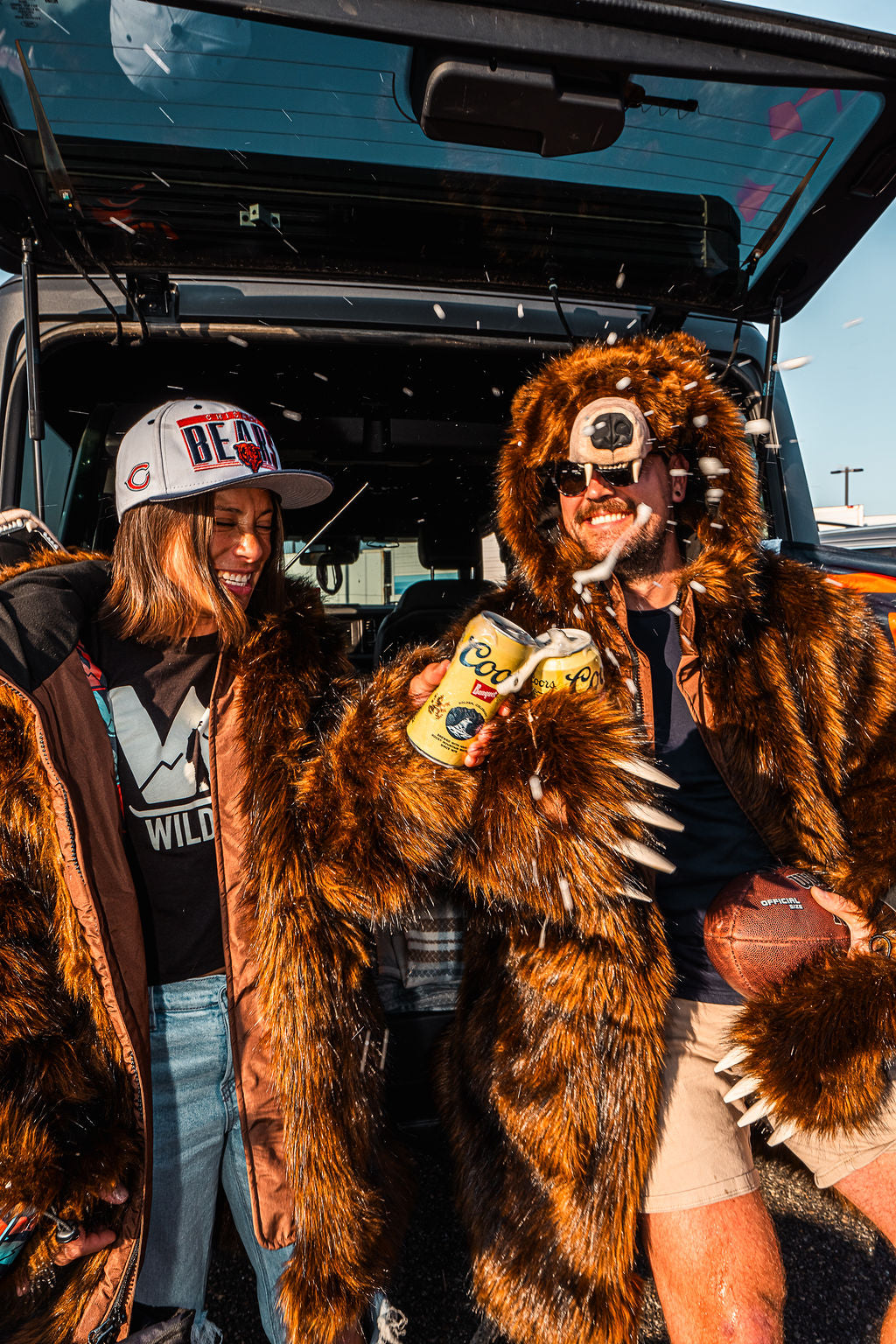 Two chicago bears fans celebrating at a tailgate by cheersing their beverages. Both are wearing wildcoat grizzly bear coats