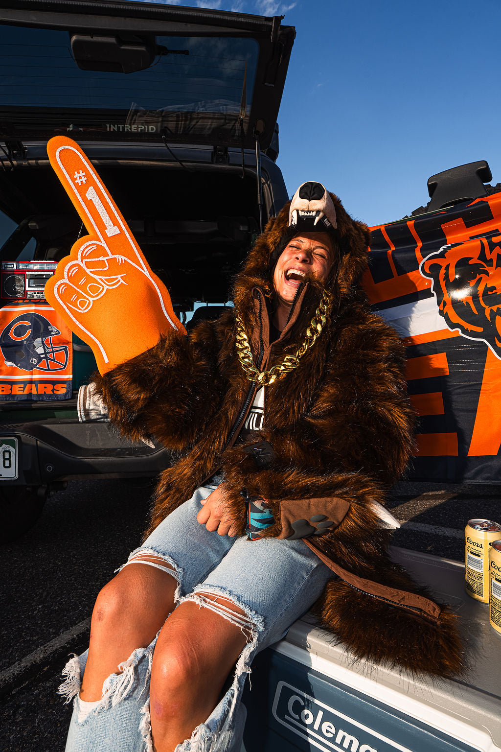 A woman wearing a brown furry costume and a bear hat sits in the back of a decorated truck, holding an orange foam finger. The truck features Chicago Bears graphics, including the team logo and 'BEARS' text, and a Coleman cooler is visible in the foreground.