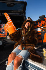 A woman wearing a brown furry costume and a bear hat sits in the back of a decorated truck, holding an orange foam finger. The truck features Chicago Bears graphics, including the team logo and 'BEARS' text, and a Coleman cooler is visible in the foreground.