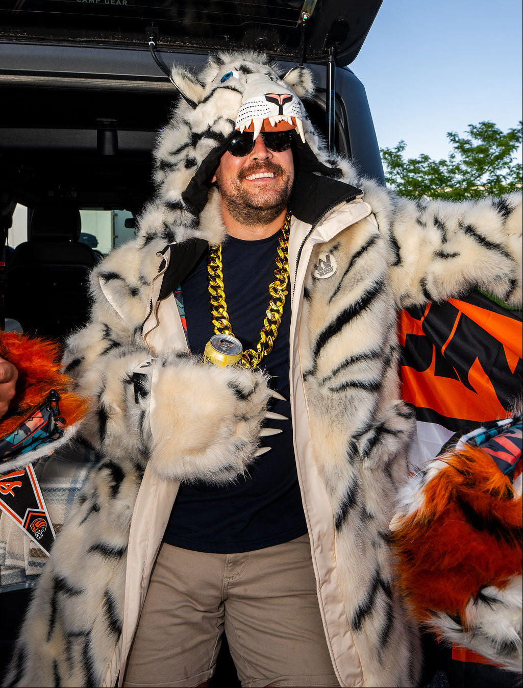 A man wearing a white faux fur tiger costume jacket with black stripes, a matching tiger mask, sunglasses, and a chunky gold chain necklace, standing in front of an open truck door.