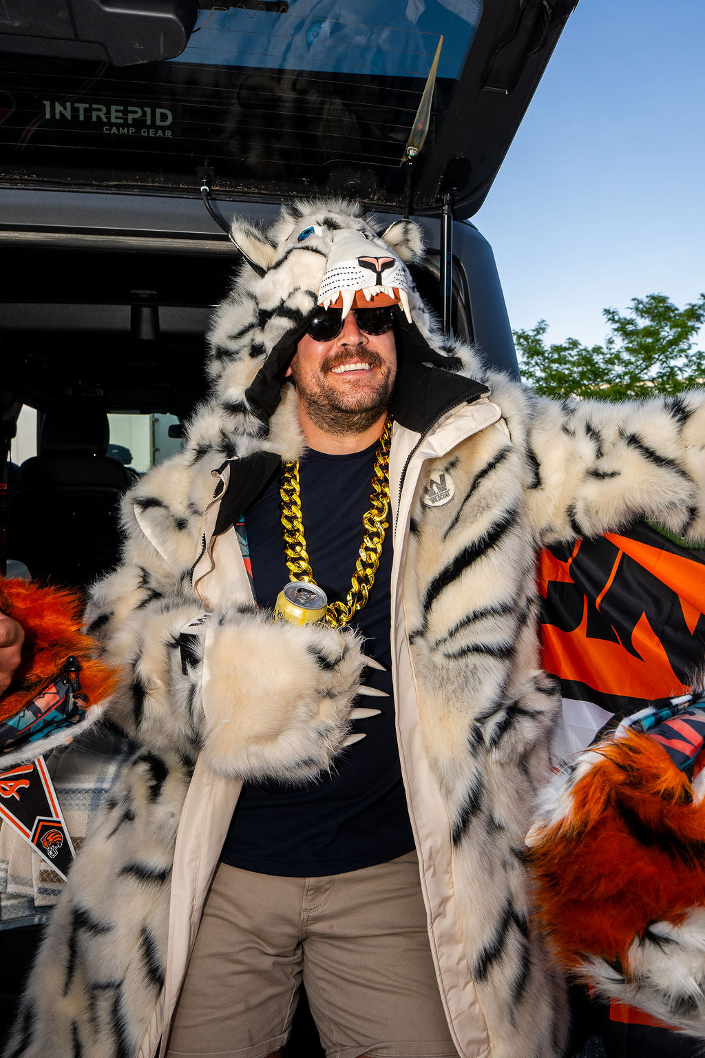 A man wearing a white faux fur tiger costume jacket with black stripes, a matching tiger mask, sunglasses, and a chunky gold chain necklace, standing in front of an open truck door.