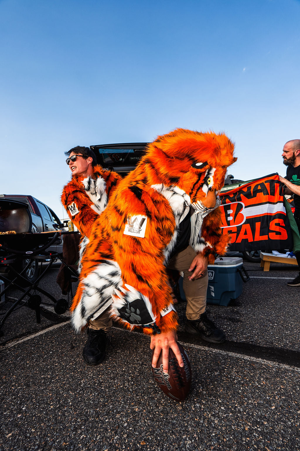 A person in a bright orange and black tiger mascot costume poses on a parking lot pavement under a clear blue sky, holding a football. Another person in a Bengals costume stands behind them holding a Bengals jersey.