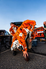 A person in a bright orange and black tiger mascot costume poses on a parking lot pavement under a clear blue sky, holding a football. Another person in a Bengals costume stands behind them holding a Bengals jersey.