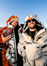 Two men wearing tiger costume hoodies and jackets, one holding a Wilson football, against a blue sky background.