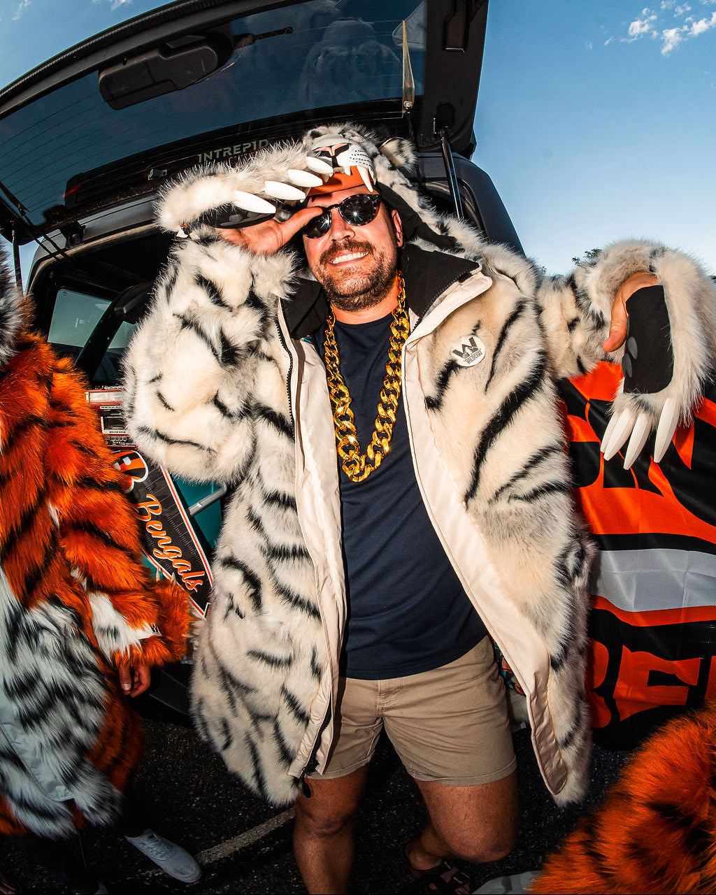A man wearing a white and black tiger-striped fur jacket, sunglasses, and a chunky gold chain stands in front of an open car trunk under a blue sky, with orange and white fur pieces visible on both sides.