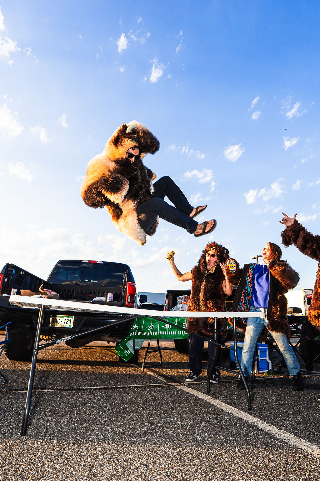 Man wearing a wildcoat great white buffalo coat jumping off the bed of a truck onto a table. Classic table smash jump at a buffalo bills tailgate. Two women are cheering as he flies through the air.
