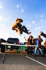 Man wearing a wildcoat great white buffalo coat jumping off the bed of a truck onto a table. Classic table smash jump at a buffalo bills tailgate. Two women are cheering as he flies through the air.