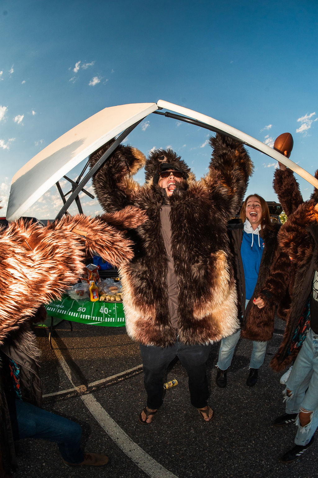 Man wearing a wildcoat great white buffalo coat holds a table over his head in celebration after successfully breaking it by jumping off a truck at a tailgate. Buffalo bills fans around him are celebrating