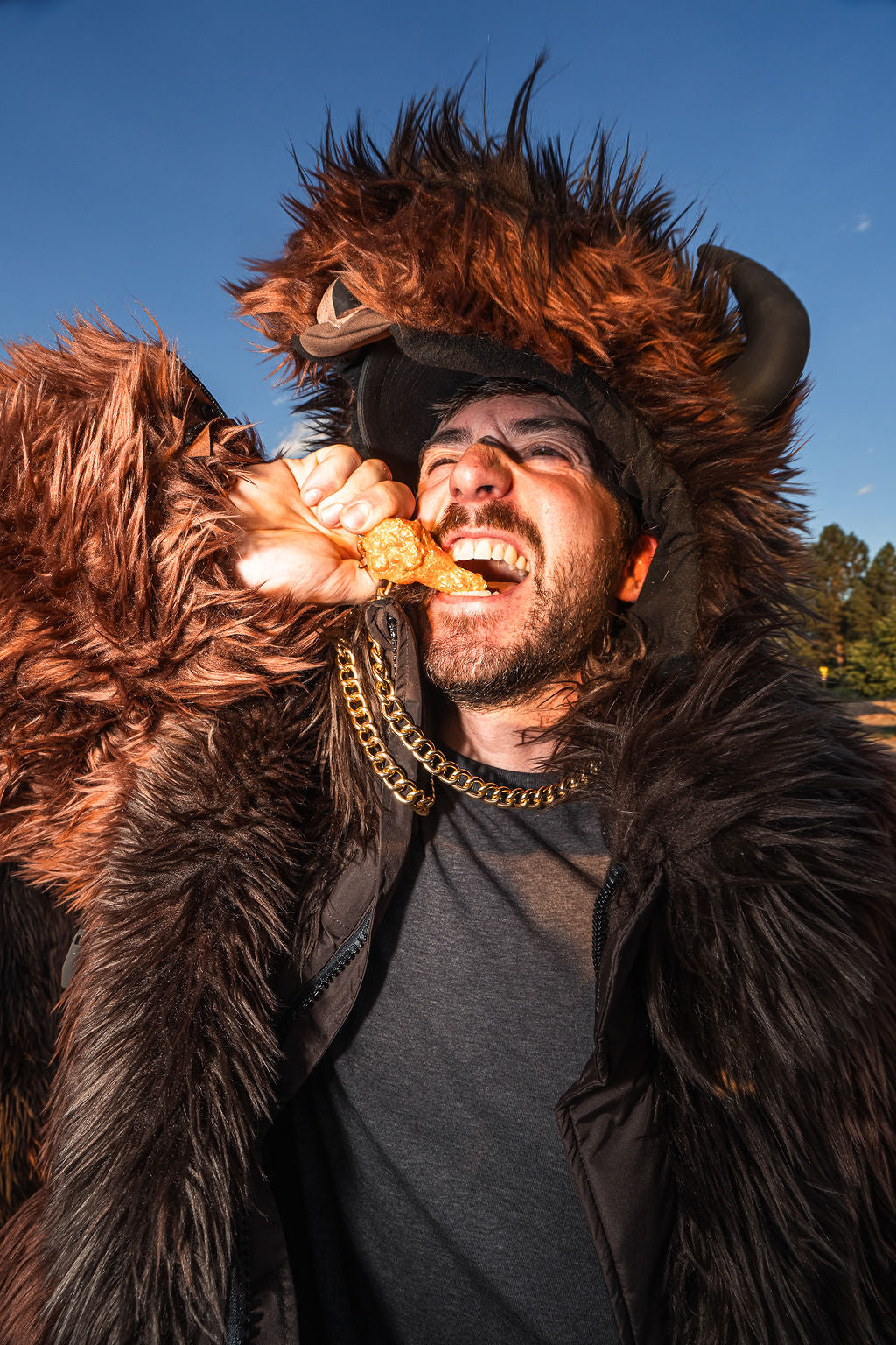 Man wearing a brown buffalo wildcoat while biting into a buffalo wing at a buffalo bills tailgate
