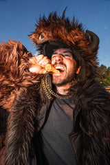 Man wearing a brown buffalo wildcoat while biting into a buffalo wing at a buffalo bills tailgate