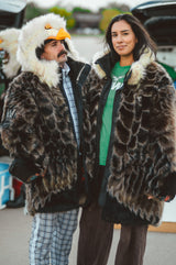 Two people standing outdoors wearing large fur coats. The person on the left wears a brown-and-white mottled eagle fur coat with a hood shaped like an eagle head with a yellow beak, over a plaid shirt and pants. The right person is also wearing a wildcoat eagle coat with the hood detached.