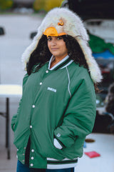 A woman wearing a green varsity-style jacket with white trim, blue jeans, and a large white faux-fur hood with a wildcoat eagle hood, standing outdoors at what looks like a tailgate or casual outdoor event.