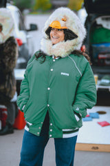 A woman wearing a green varsity-style jacket with white trim, blue jeans, and a large white faux-fur hood with a wildcoat eagle hood, standing outdoors at what looks like a tailgate or casual outdoor event.
