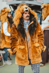 A man wearing a plush lion costume jacket with a large tan and brown faux-fur hood, dark mane details, and a matching lion headpiece, standing outdoors with other people blurred in the background of a detroit lions tailgate.