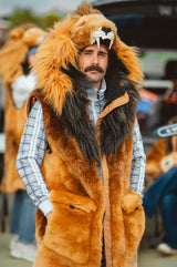 A man wearing a lion costume vest with a plush orange and black mane hood, layered over a plaid shirt, photographed outdoors at what looks like a street fair or event.