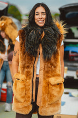 A woman wearing a long, plush lion-face fur vest in warm golden brown and black, standing outdoors at what looks like a market or event.