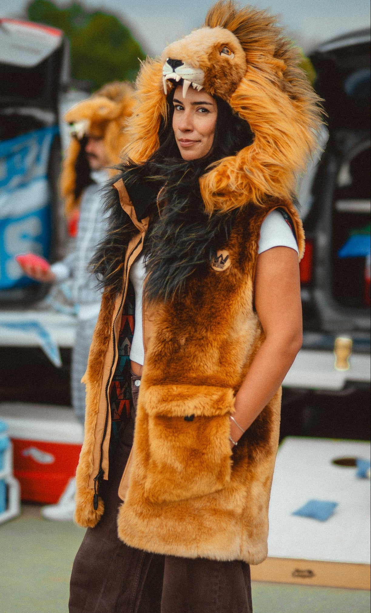 A woman wearing a plush lion costume vest with a large furry hood featuring a lion face and mane, standing outdoors near vehicles and coolers at a detroit lions tailgate