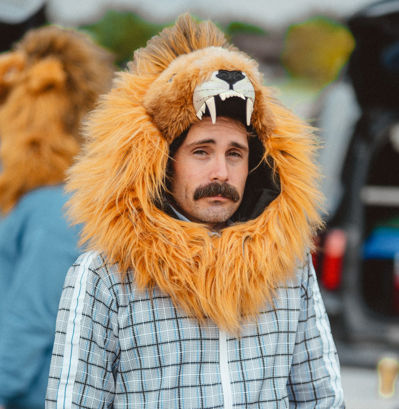 A man wearing a gray and white plaid zip-up jacket with white stripes on the sleeves, matching plaid pants, and a large orange faux-fur lion hood with a lion face mask.