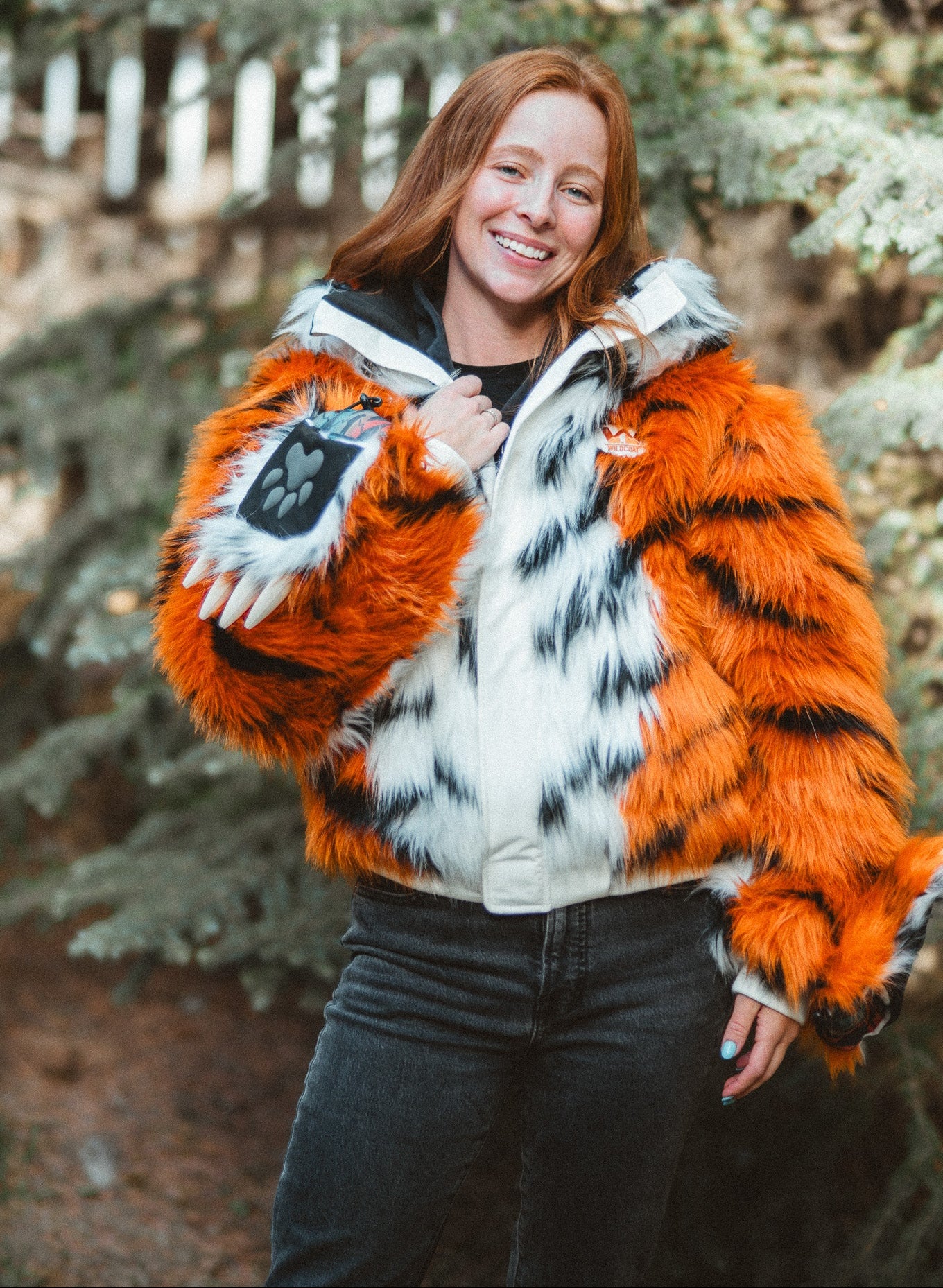 A woman wearing a hooded faux fur jacket with a tiger-stripe pattern in orange, black, and white, standing outdoors in front of blurred trees.