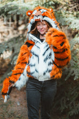 A woman wearing a plush orange and black tiger costume jacket with a matching hood featuring a tiger face, standing outdoors in front of blurred trees.