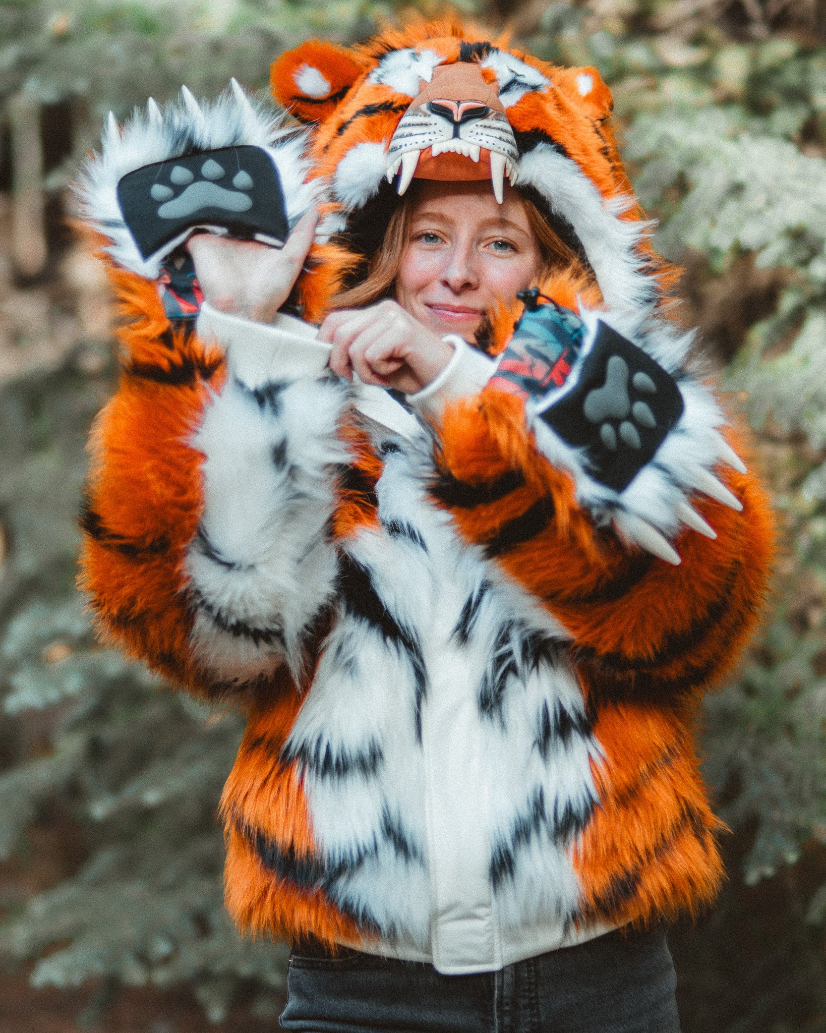 A person wearing a plush tiger costume jacket with orange, black, and white fur-like patterning, a tiger hood with animal ears and snout, and paw-print hand covers, photographed outdoors against blurred greenery.