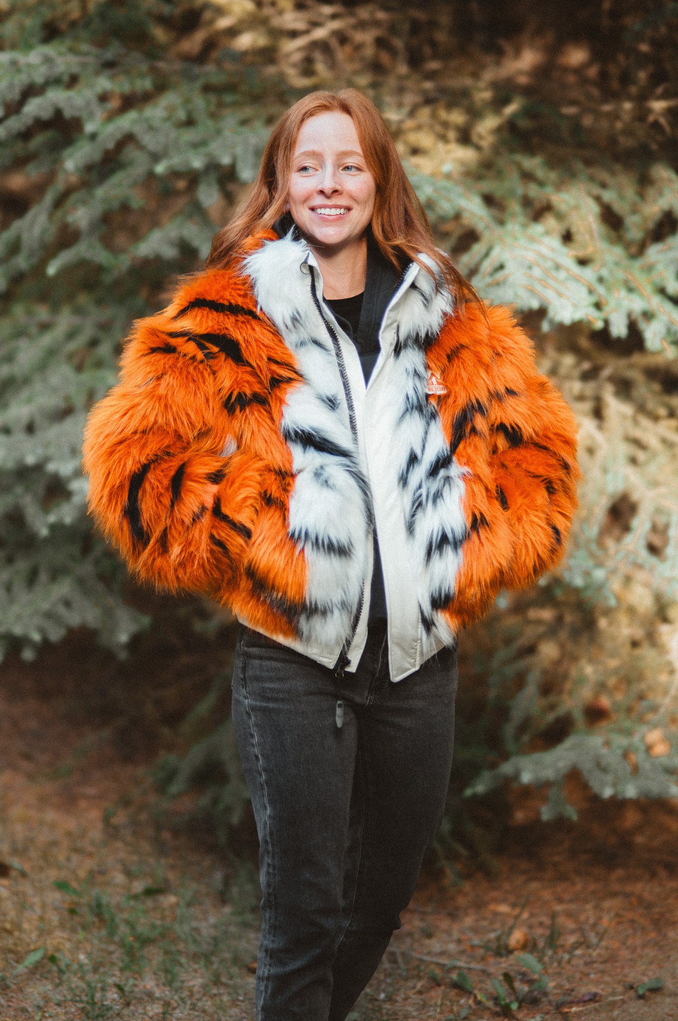 A woman wearing a vibrant orange and black faux fur tiger patterned winter jacket with a white and black patterned fur collar, standing outdoors in front of a blurred forest background.