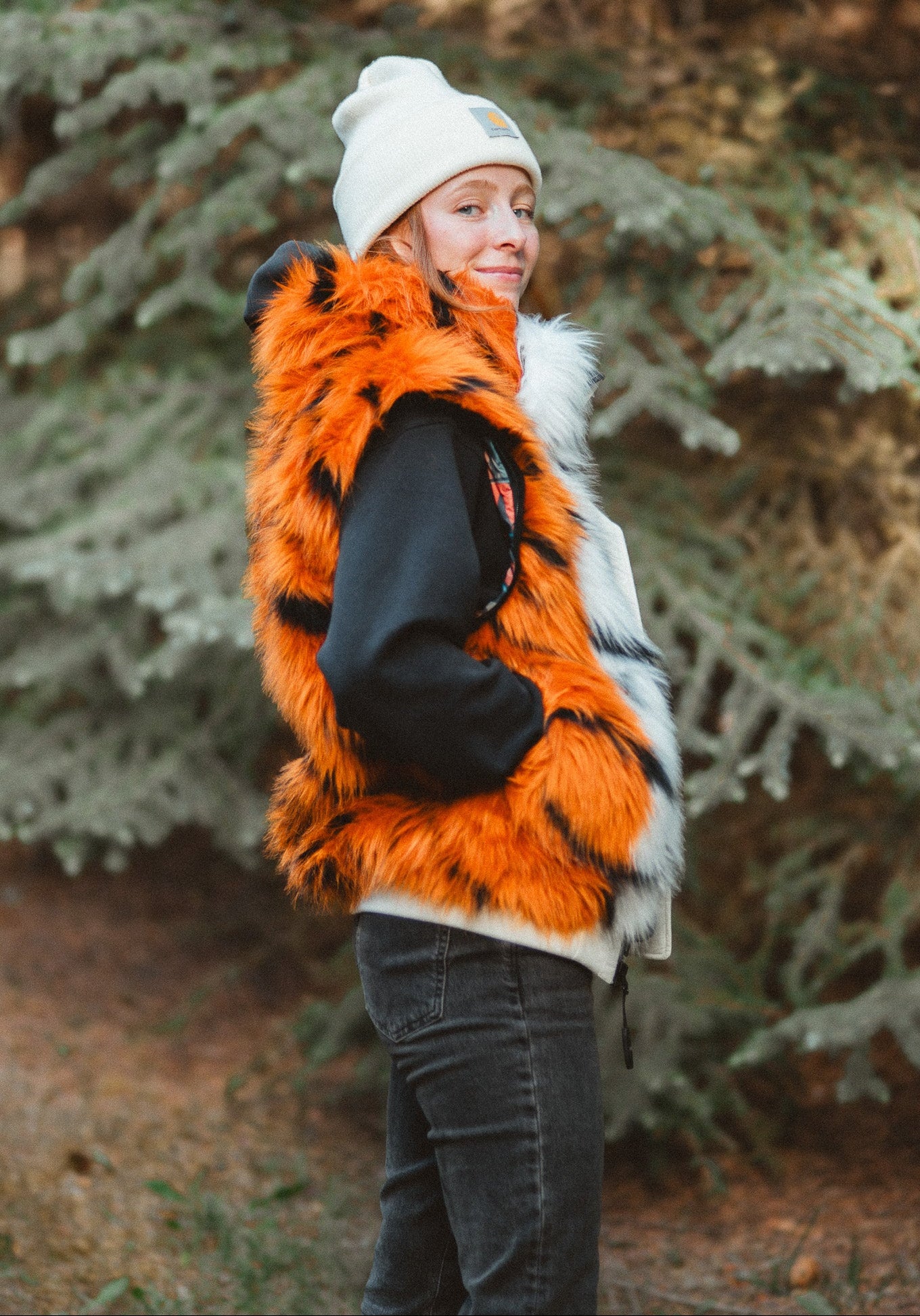 A woman wearing a Wildcoat tiger bomber jacket, bright orange faux fur vest with black tiger-like stripes, a black jacket, a white beanie, and dark jeans, standing outdoors in front of a blurred evergreen tree.