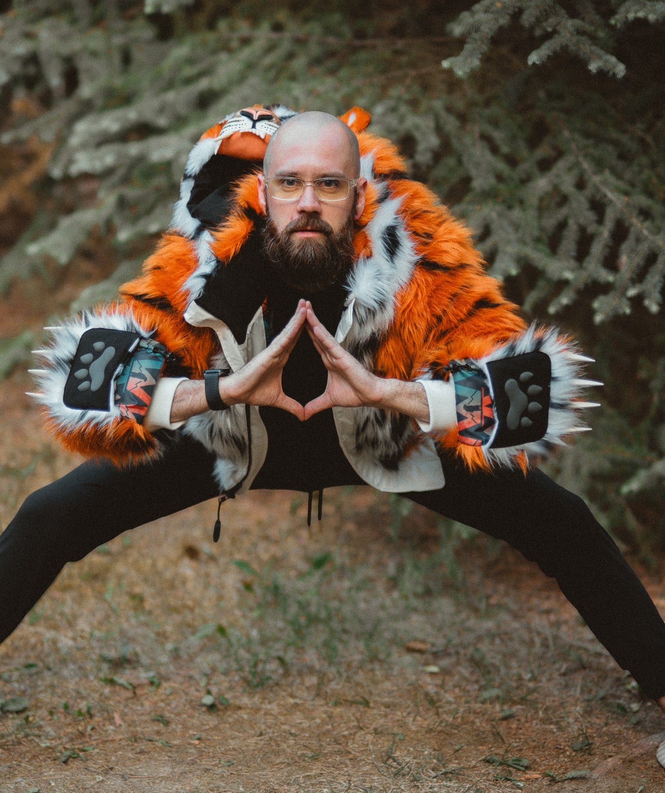 A man wearing the wildcoat orange tiger bomber coat in a forest pose. The coat is a bright orange and white tiger-print faux fur jacket with black and white striped hood details, black pants, and clawed mittens flipped back.