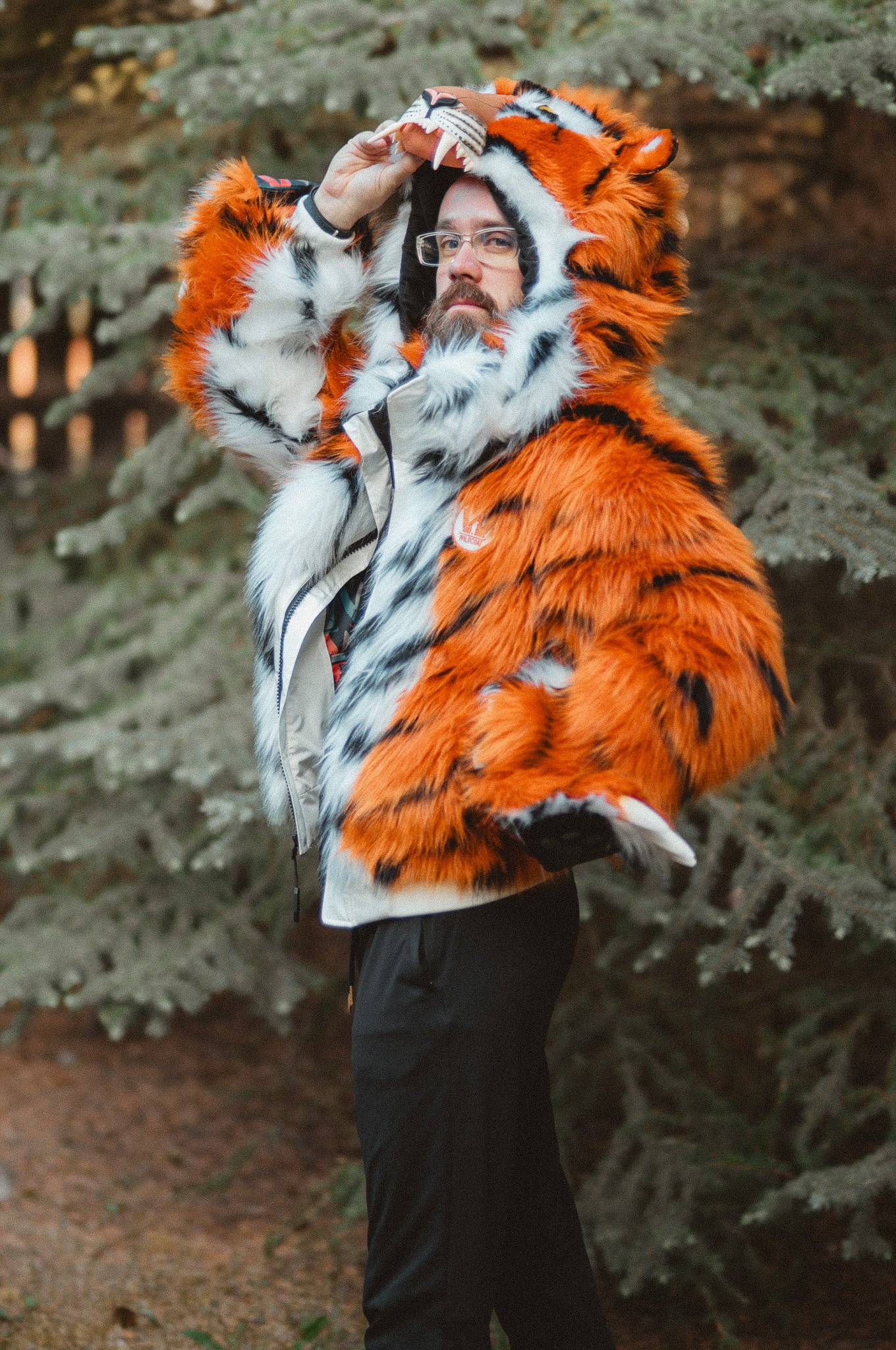 A person wearing a plush tiger-print hooded jacket with orange, black, and white fur-like texture, standing outdoors in front of evergreen trees.