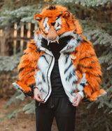 A person wearing a plush tiger costume jacket with a matching tiger head mask, standing outdoors in front of evergreen trees. The costume features bright orange faux fur with black stripes and white panels.