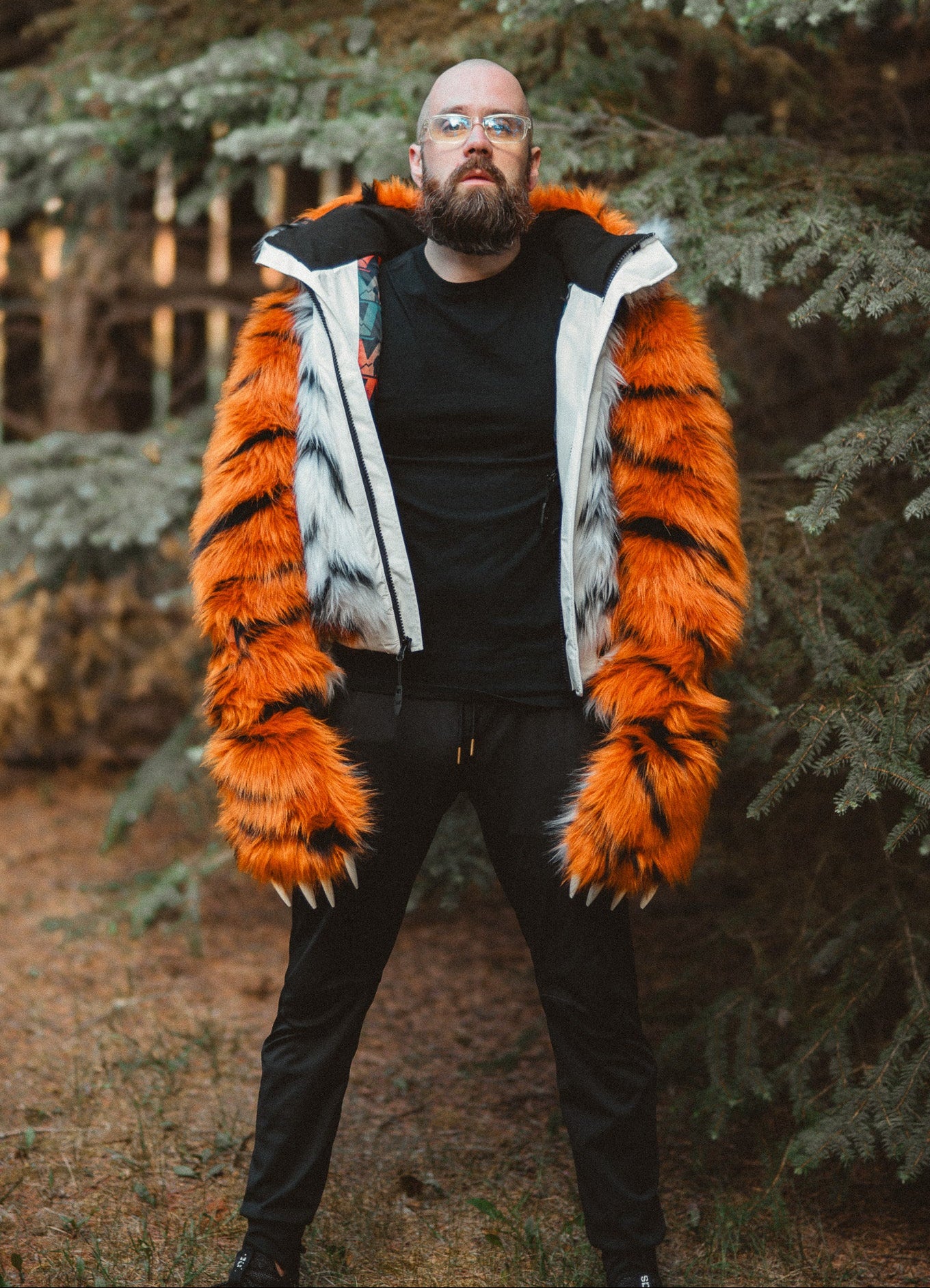 A man wearing a black t-shirt and black pants stands outdoors in front of evergreen trees, wearing a unique orange and white faux fur jacket with black tiger-like stripes.