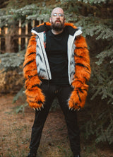 A man wearing a black t-shirt and black pants stands outdoors in front of evergreen trees, wearing a unique orange and white faux fur jacket with black tiger-like stripes.
