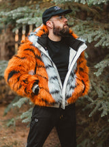 A man wearing the Wildcoat orange tiger bomber coat, a vibrant orange and black faux fur jacket with white lining, a black t-shirt, black pants, and a black baseball cap, standing outdoors in a forest setting.