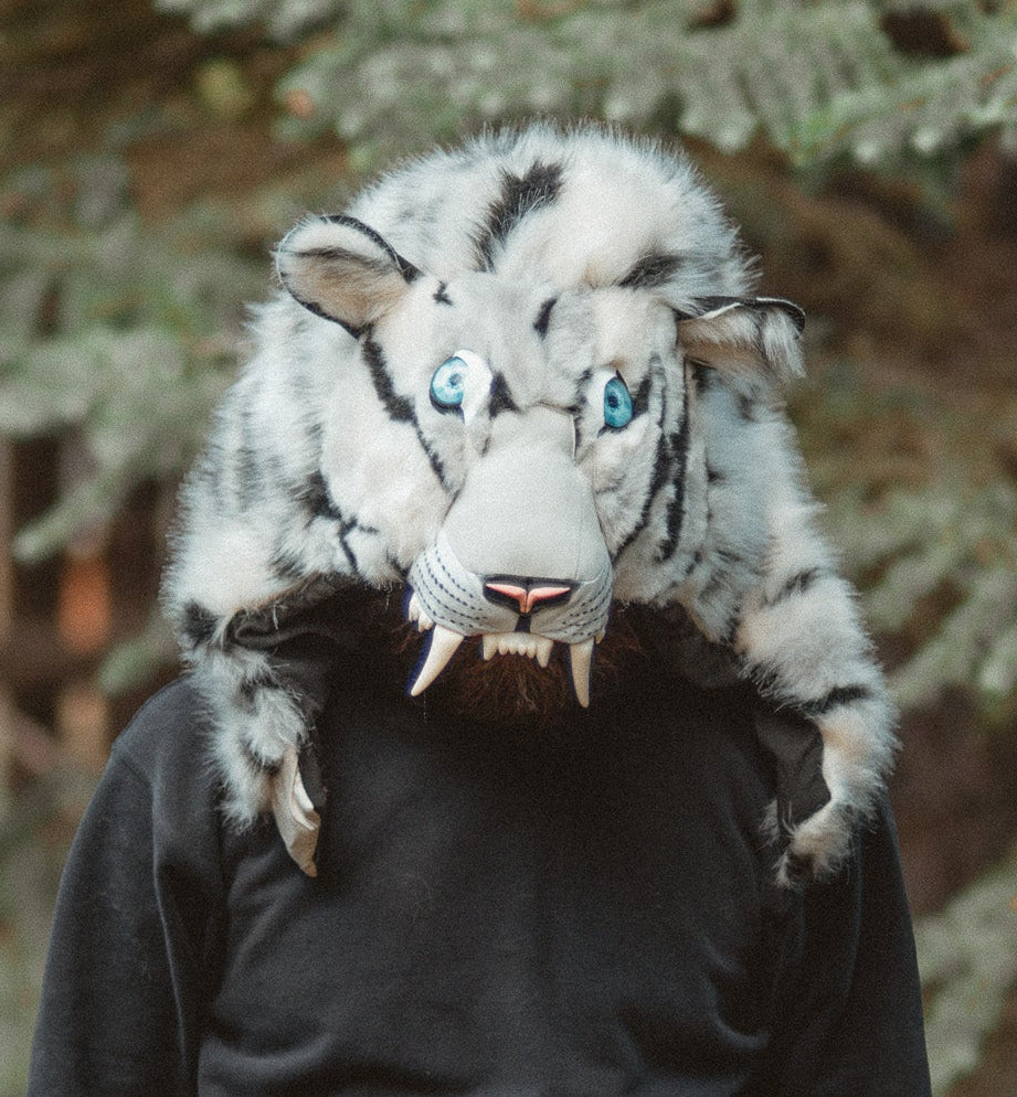 A person wearing a black hoodie and black sweatpants stands outdoors in front of evergreen trees, wearing a large white and gray tiger mask with blue eyes and fangs over the head.
