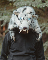 A person wearing a black hoodie and black sweatpants stands outdoors in front of evergreen trees, wearing a large white and gray tiger mask with blue eyes and fangs over the head.