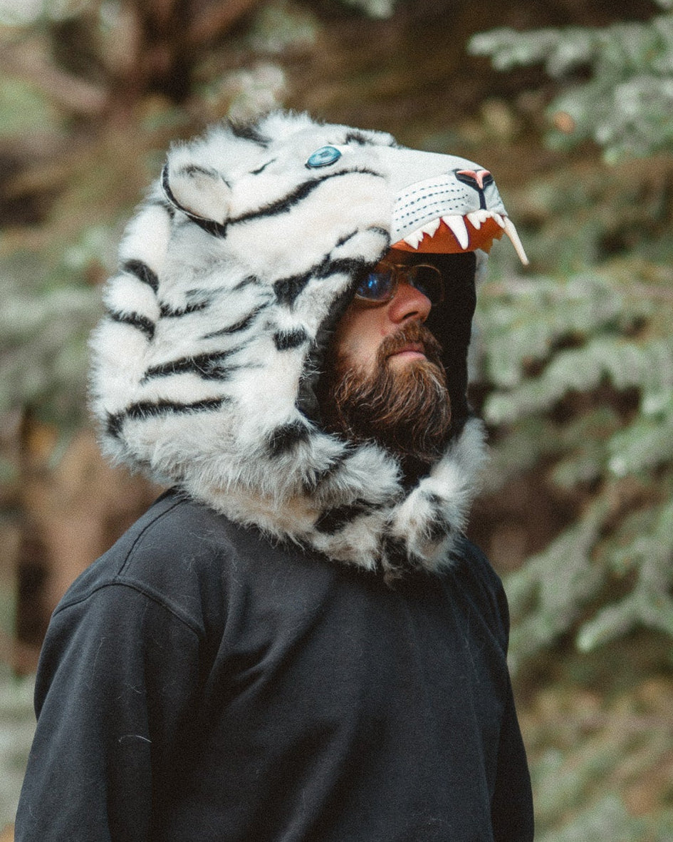 A man wearing a black hoodie and black sweatpants stands outdoors in a forest, wearing a plush white tiger-face hood with black stripes and an open mouth showing teeth.