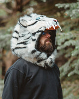 A man wearing a black hoodie and black sweatpants stands outdoors in a forest, wearing a plush white tiger-face hood with black stripes and an open mouth showing teeth.