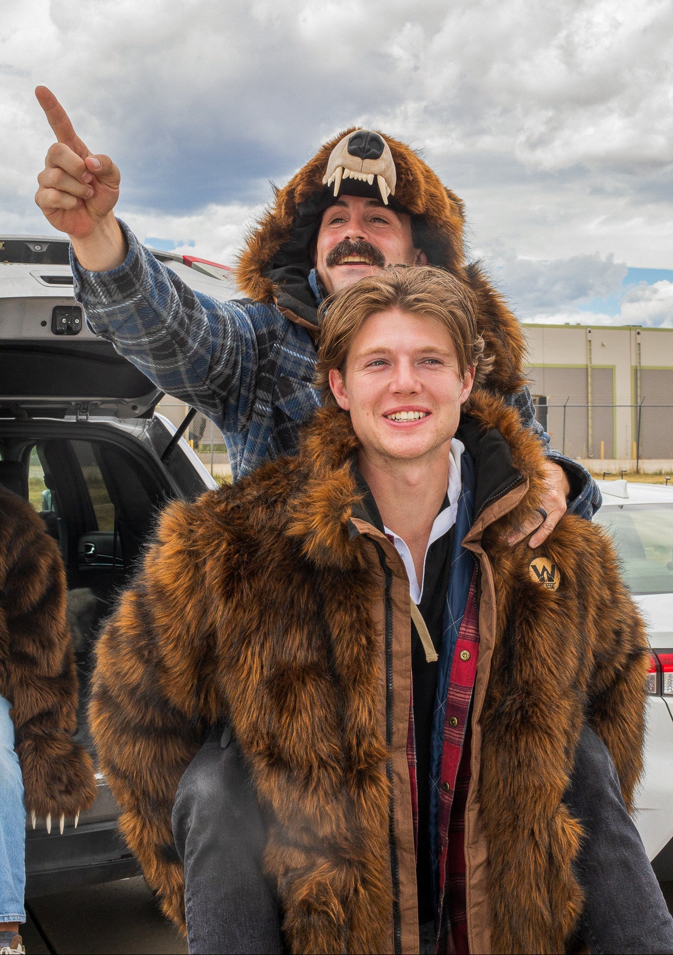 Two men at a bears tailgate wearing wildcoat grizzly bear parkas. one man is giving the other a piggyback ride, the man on his back is wearing the grizzly bear hood from the first mans coat