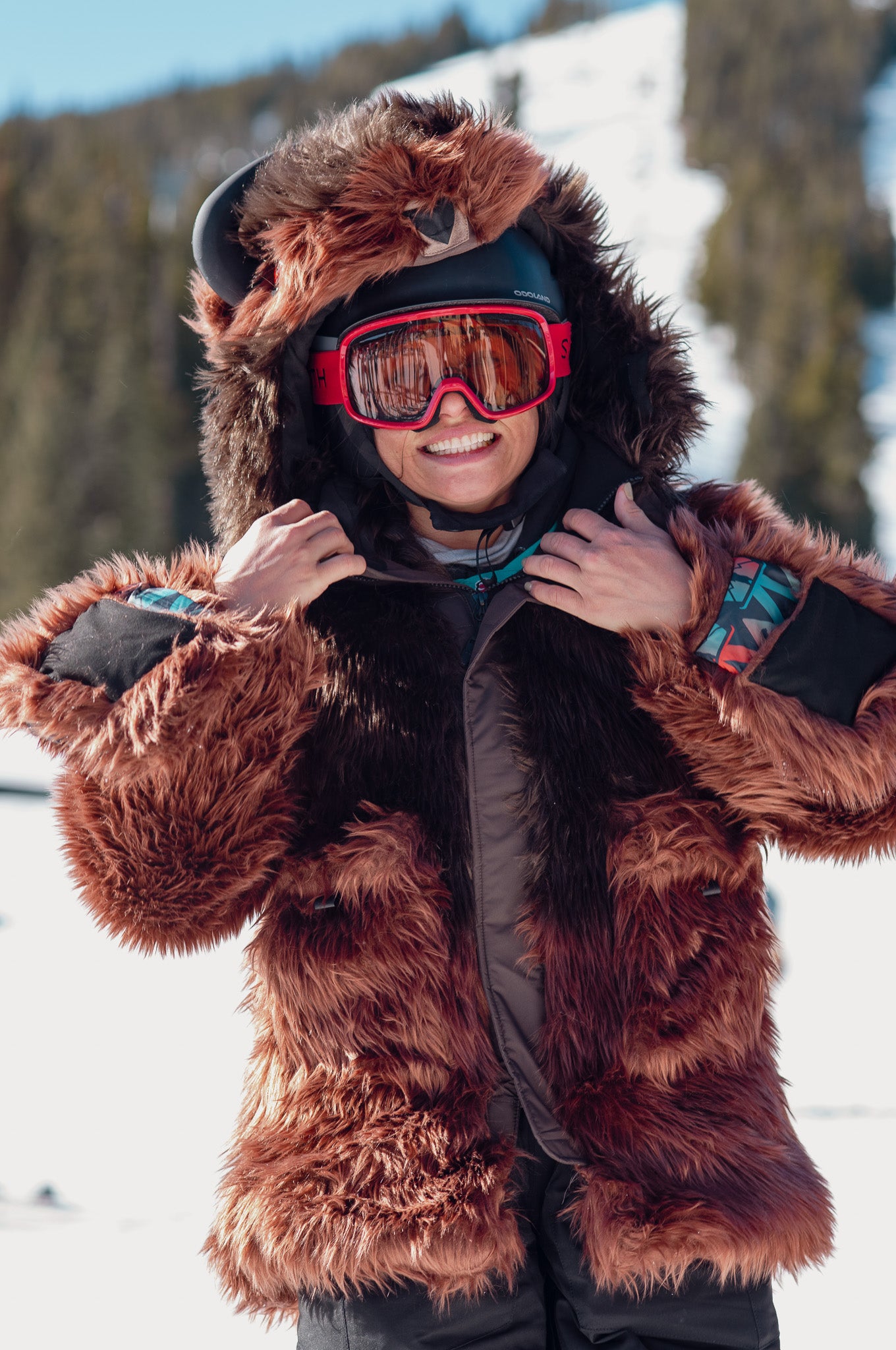 woman wearing a wildcoat buffalo bison coat and smiling at a ski resort. the horned hood of the bison is over her ski helmet