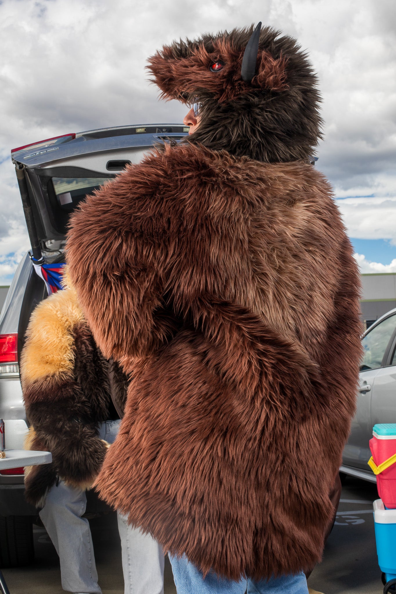 woman wearing a wildcoat v6 buffalo bison faux fur winter coat at a tailgate  showing the brown and gold paattern on the back of the coat with a man in the background wearing the great white buffalo coat